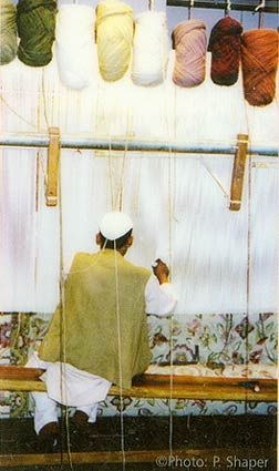 photo depicting the fine art of hand knotting a wool rug as a worker is engaged in the process of weaving a large vertical rug loom - India
